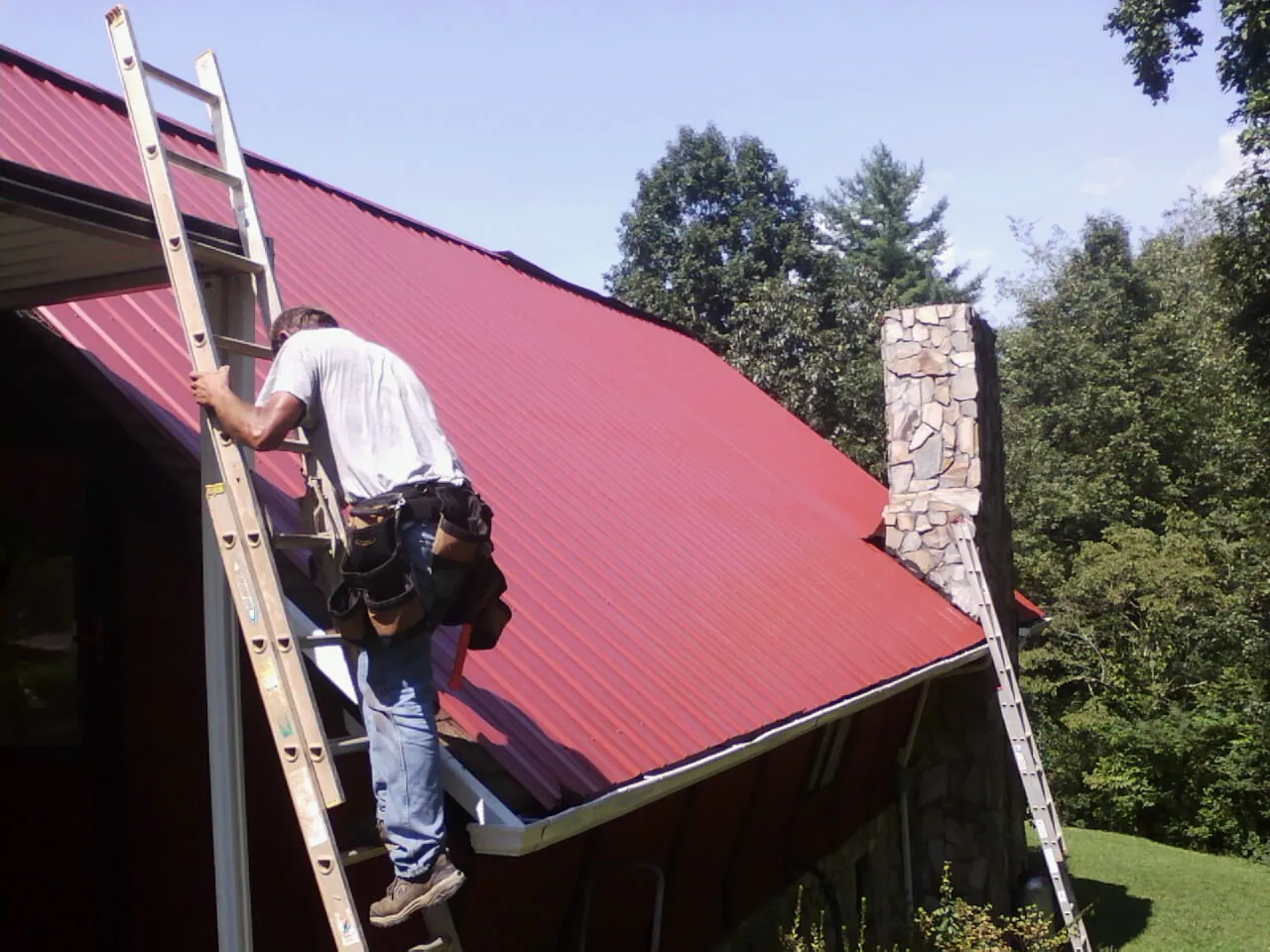 Roofer on a ladder beside a red metal roof and stone chimney — Soffit & Fascia Repair by Roofing Brothers in Berrien Springs